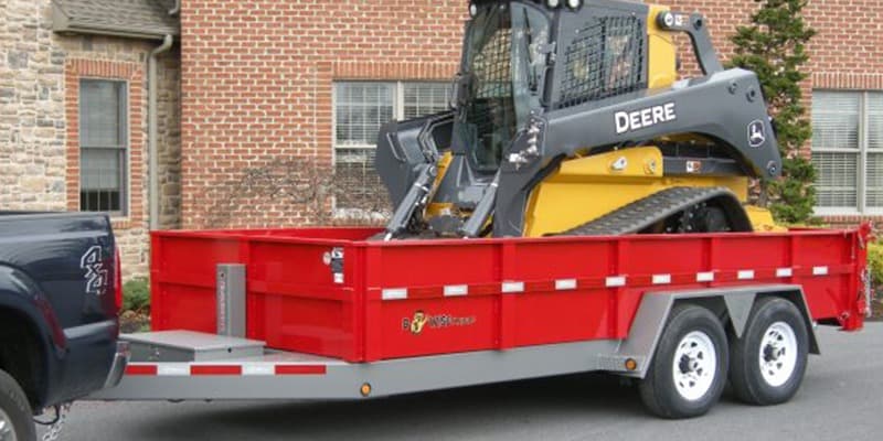 Truck hauling red B-Wise utility trailer with John Deere Skid steer in cargo